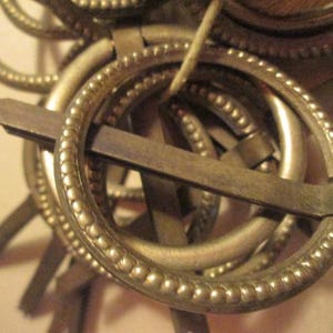 May include: A close-up of a gold-toned metal belt buckle with a decorative beaded edge. The buckle is made up of multiple interlocking rings, creating a complex and intricate design.