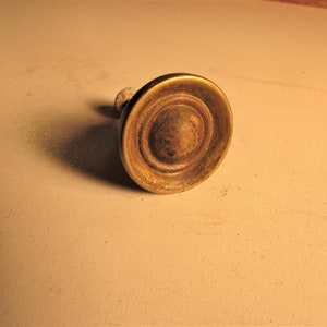 May include: A vintage brass cabinet knob with a circular, ridged design. The knob has a warm, aged patina, suggesting it is an antique or a reproduction. The knob is mounted on a small, light-colored wooden stem.
