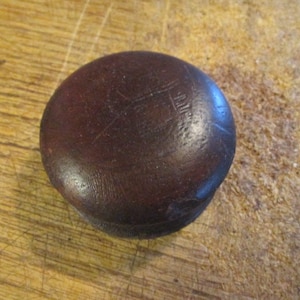 May include: A dark brown, rounded wooden knob with a slightly domed top. The knob has a smooth, polished surface with subtle variations in color and texture. It is set against a light brown, wooden background.
