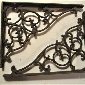 May include: Two antique, dark brown cast iron shelf brackets. Each bracket features an ornate, scrolling design with curved lines and circular elements. The brackets are rectangular with a flat base and a vertical support.
