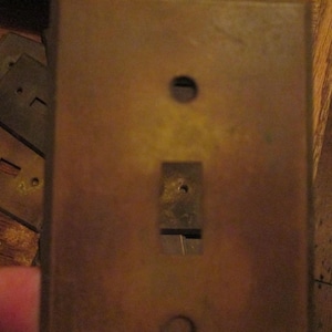 May include: A rectangular, weathered brass light switch wall plate with a dark, tarnished patina. The plate features a central rectangular cutout for a toggle switch and two circular screw holes positioned at the top and bottom of the plate.
