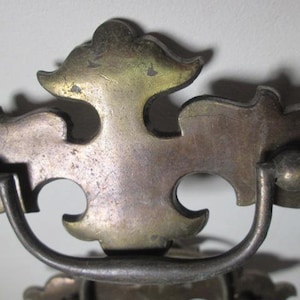 May include: Close-up of antique metal drawer pulls. The pulls have a decorative, ornate design with a curved handle and a tarnished, aged appearance. The metal has a mix of silver and gold tones.