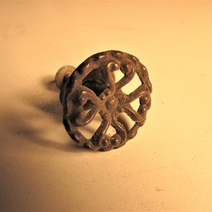 May include: Ornate, antique bronze cabinet knob with a circular, openwork design. The knob features a detailed, symmetrical pattern with a central hub and radiating, curved elements. The hardware is likely for furniture.