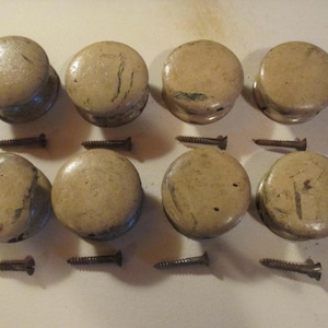 May include: A set of eight round, beige-colored cabinet knobs with a smooth, slightly weathered finish. Each knob is accompanied by a single metal screw, arranged in two rows of four, displaying a uniform circular shape and a neutral tone.