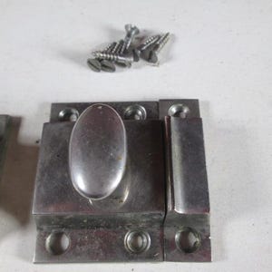May include: Close-up of a set of silver-colored metal latches with an oval handle and matching screws. The latches are rectangular with rounded edges and circular holes for mounting.