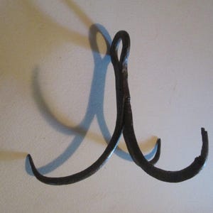 May include: A dark gray, hand-forged metal hook featuring a central vertical stem that splits into two curved prongs at the bottom. The top of the stem forms a circular loop for mounting. The metal surface has a textured, uneven, and rustic finish.