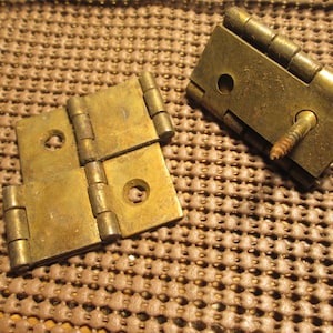 May include: Two brass-colored metal hinges with a weathered finish. One hinge is folded flat with two visible screw holes, while the other hinge is partially open and features a single metal screw protruding from one of the mounting plates.