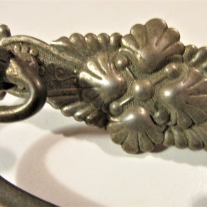 May include: Close-up of an antique silver-toned drawer pull with ornate floral and leaf detailing. The handle curves gracefully, and the decorative elements are raised, creating a textured surface. A vintage hardware piece.