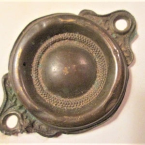 May include: Antique brass door knob with a circular design. The knob features a central, raised circular element surrounded by a textured ring. The edges have decorative flourishes and mounting holes. The brass has a weathered patina.