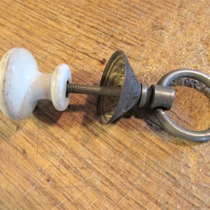 May include: An antique door pull with a white ceramic knob, a decorative metal cone, and a silver ring. The knob has a slightly aged appearance. The hardware is designed for a door or drawer.