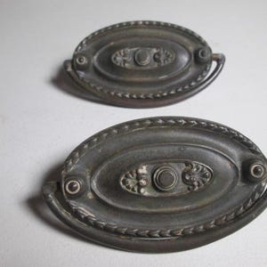 May include: Two antique bronze oval drawer pulls. Each pull features a central circular design with floral accents and a small handle. The pulls have a decorative rope-like border and are set against a white background.