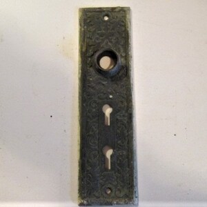 May include: An antique, rectangular door plate with an ornate floral design. The plate is dark gray with a circular hole at the top and two keyholes below. The edges show signs of wear.