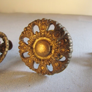 May include: Close-up of three antique brass drawer pulls. The center pull is a detailed, circular design with a central disc and ornate floral-like patterns. The brass has a warm, aged patina, suggesting vintage or repurposed use.