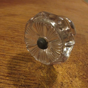May include: A clear glass door knob with a faceted design and a dark center. The knob has a circular shape with a ridged pattern radiating from the center. The glass is transparent, allowing light to pass through.