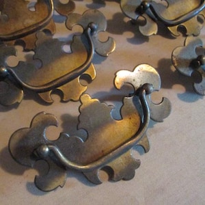 May include: Antique brass drawer pulls with a decorative, ornate design. Each pull features a curved handle and a stylized, floral-like backplate. The hardware has a vintage, aged appearance, suggesting use in older furniture.