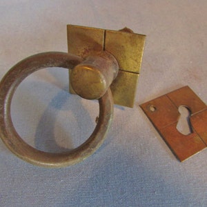 May include: Antique brass door pull with a ring handle and square backplate. A separate brass keyhole cover is also present. The hardware has a weathered appearance, suggesting age and use. Ideal for vintage or rustic decor.