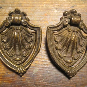 May include: Two antique brass drawer pulls. Each pull has a shield shape with a detailed shell design in the center. The pulls have a decorative top and a curved handle. The brass has a warm, aged patina.