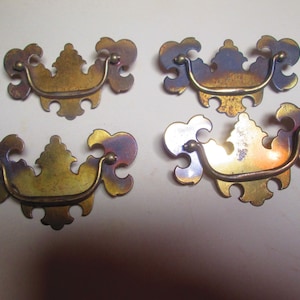 May include: Four antique brass drawer pulls with a decorative, ornate design. Each pull features a curved handle and a detailed backplate with a mix of gold, bronze, and purple hues. These vintage hardware pieces are ideal for furniture restoration projects.