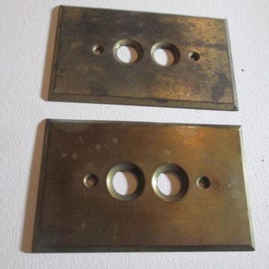 May include: Two rectangular, aged brass electrical switch plates. Each plate has two circular holes and three smaller screw holes. The brass has a tarnished, vintage appearance.