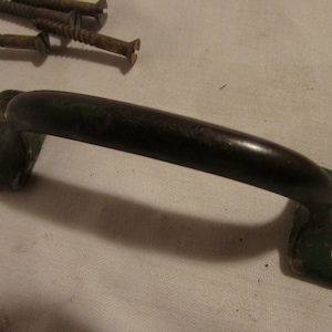 May include: An antique cabinet handle with a dark brown, curved grip and green mounting plates. Three rusty screws are visible in the background. The handle has a vintage, worn appearance.