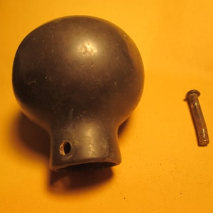 May include: A dark, matte, spherical knob with a cylindrical base featuring a small circular hole on the side. Beside the knob lies a separate, slender, metallic pin with a flat head, showing signs of oxidation and a weathered, brownish texture.