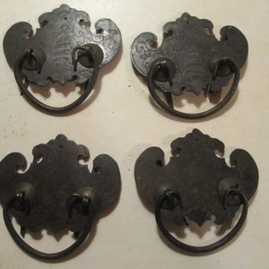 May include: Four antique black metal drawer pulls with ornate, shield-like backplates and circular handles. The pulls have a decorative design etched into the metal, adding a vintage aesthetic. These hardware pieces are ideal for furniture restoration projects.