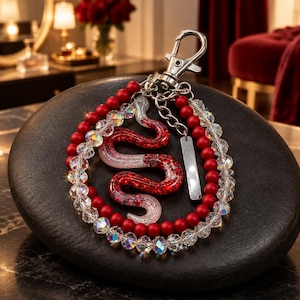 May include: A keychain featuring a red and white glittery snake charm, matte red beads, and clear faceted crystal beads. The charm is attached to a silver-toned metal lobster clasp and a small rectangular metal tag, creating a decorative accessory.