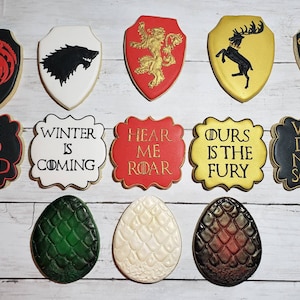 May include: A collection of decorated sugar cookies with various designs and text. The cookies feature shield shapes with house sigils and phrases like "Fire and Blood" and "Winter is Coming." Also included are three dragon egg cookies.