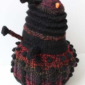 May include: A black and red knitted toy with a round body and a pointed top. The toy has a textured surface and small orange pom-poms on the top.