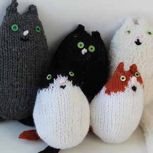 Könnte beinhalten: Fünf handgestrickte Katzen-Plüschtiere in verschiedenen Farben. Die Katzen haben Knopfaugen, kleine Nasen und einfache Lächeln. Zu den Farben gehören Grau, Schwarz, Weiß sowie Orange und Weiß. Die Spielzeuge sind weich und kuschelig.
