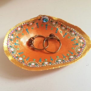 Könnte beinhalten: Eine kleine orangefarbene Muschelschalen-Schale mit goldenen Verzierungen und weißen, blauen und grünen Punkten. Zwei goldene Ringe liegen in der Schale.