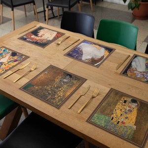 Puede incluir: Seis manteles individuales cuadrados con un borde dorado y una imagen impresa de la pintura "El Beso" de Gustav Klimt. Los manteles individuales están sobre una mesa de madera con cubiertos de oro.