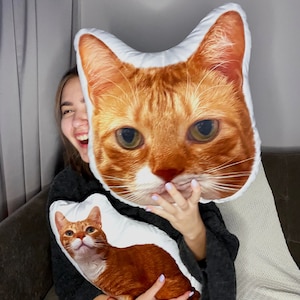 Op de afbeelding: Een vrouw houdt twee kussens in de vorm van een kat vast. Het ene kussen is een close-up van een kattenkop, het andere is een afbeelding van een kat in zijn geheel.