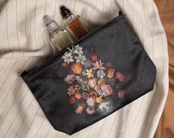 Op de afbeelding: Zwarte fluwelen toilettas met een bloemenprint met een boeket kleurrijke bloemen. Twee glazen flessen met zilveren en zwarte doppen zitten in de tas.