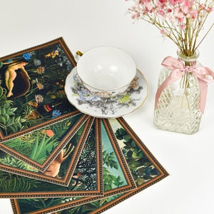 Puede incluir: Una taza y un platillo blancos con dise&ntilde;os florales se asientan sobre una pila de posavasos decorativos con escenas de la jungla. Tambi&eacute;n hay un peque&ntilde;o jarr&oacute;n de cristal con flores rosas y una cinta rosa.