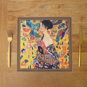 May include: A square placemat with a gold frame and a colorful painting of a woman with a peacock feather fan. The painting is set against a yellow background with flowers and birds.