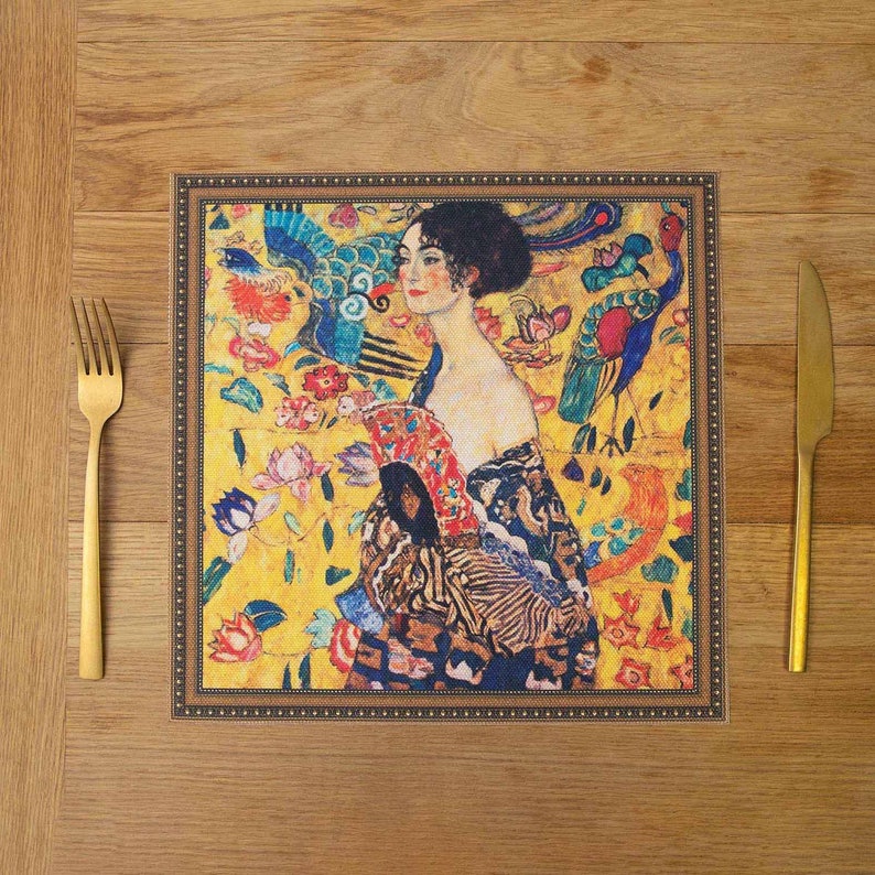 May include: A square placemat with a gold frame and a colorful painting of a woman with a peacock feather fan. The painting is set against a yellow background with flowers and birds.