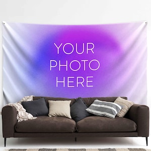 Pode incluir: Uma tapeçaria de parede retangular com um fundo gradiente roxo e azul vibrante com o texto branco YOUR PHOTO HERE centralizado no meio, posicionado acima de um sofá marrom escuro com várias almofadas decorativas.