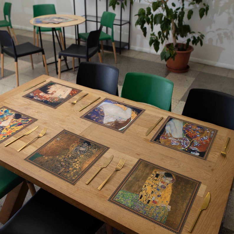 May include: Six square placemats with different images of famous paintings by Gustav Klimt. The placemats are brown with a gold border and are laid out on a wooden table.