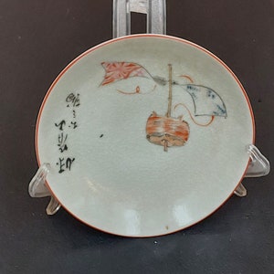 Puede incluir: Un pequeño plato de porcelana blanca con un borde rojo. El plato presenta un diseño pintado a mano de una cometa roja y blanca con una cometa azul y blanca volando detrás de ella. El plato también tiene caligrafía negra.