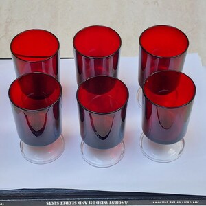 May include: Six vintage red-tinted glass goblets with clear stems are arranged on a white surface. The glasses have a cylindrical shape and a deep red colour, with a clear base. The goblets are likely for serving beverages.