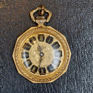 Könnte beinhalten: Eine goldfarbene Taschenuhr mit schwarzem Zifferblatt und römischen Ziffern für die Stunden. Die Uhr hat ein dekoratives Design am Rand und eine kleine Krone oben. Das Zifferblatt der Uhr zeigt "Rexer Swiss Made".