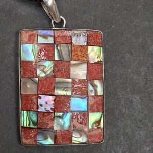 May include: Rectangular pendant with a mosaic design. The pendant features a checkerboard pattern of red and iridescent abalone shell squares, set in a silver-tone frame. A black cord is attached to the pendant.