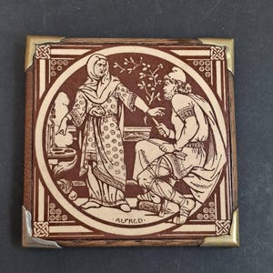 Puede incluir: Un azulejo cuadrado de madera con una ilustración en marrón y crema de una persona con pañuelo en la cabeza señalando a una figura sentada que sostiene un arco y una flecha. La palabra ALFRED está impresa abajo. Tiene esquineros de metal dorado.