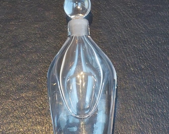 Puede incluir: Un frasco de perfume de vidrio transparente con un tapón redondeado. La botella tiene una forma única con estrías verticales y una base acampanada. El tapón es una pequeña esfera transparente. La botella está vacía.