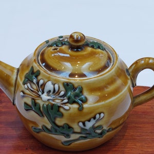 May include: A small, brown ceramic teapot with a floral design. The teapot has a lid and a handle. It is sitting on a wooden base.