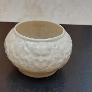 May include: Cream-coloured, round ceramic bowl with a detailed relief pattern. The pattern features faces and grapevines. The bowl has a wide opening and a slightly raised base. A decorative piece for the home.