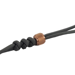 May include: A close-up of a dark gray paracord with a copper-colored bead. The bead has a textured surface and is knotted into the paracord.