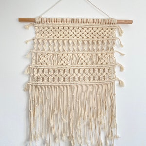 Peut inclure: Une suspension murale en macramé beige avec un motif géométrique et des franges. La suspension est faite de cordon de coton naturel et est fixée à une baguette en bois.