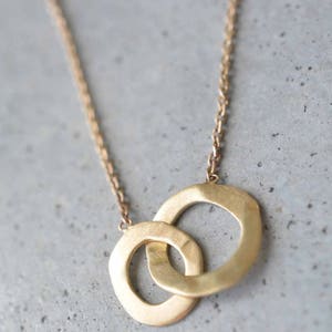 May include: A gold necklace with a pendant featuring two interlocking circles. The circles are smooth and have a slightly hammered texture.